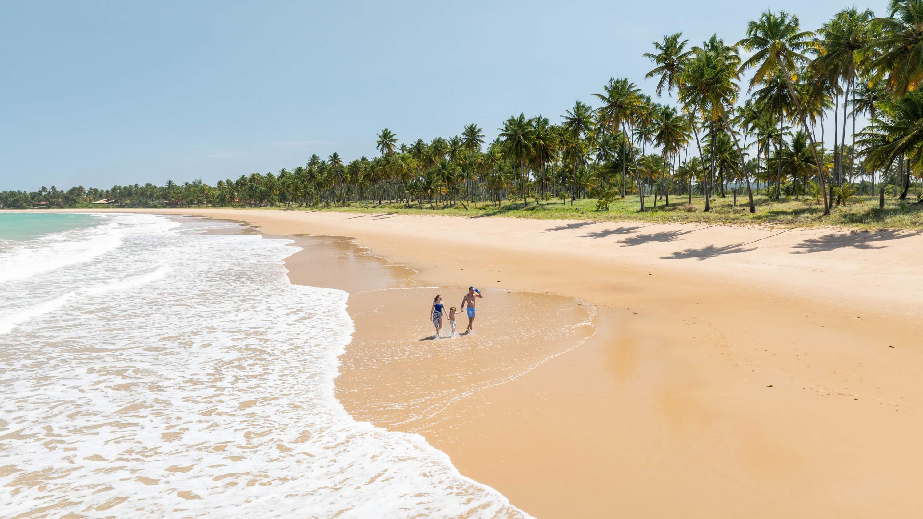 Família feliz caminhando pela faixa de areia entre o mar e a densa vegetação de coqueiros da praia de Ipioca, em Alagoas.